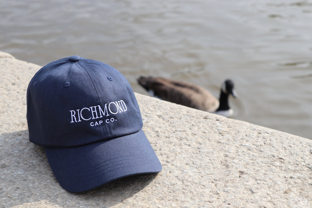 A navy blue baseball cap with 'RICHMOND CAP CO.' embroidered in white on the front, resting on a stone ledge with a duck in the water behind it.