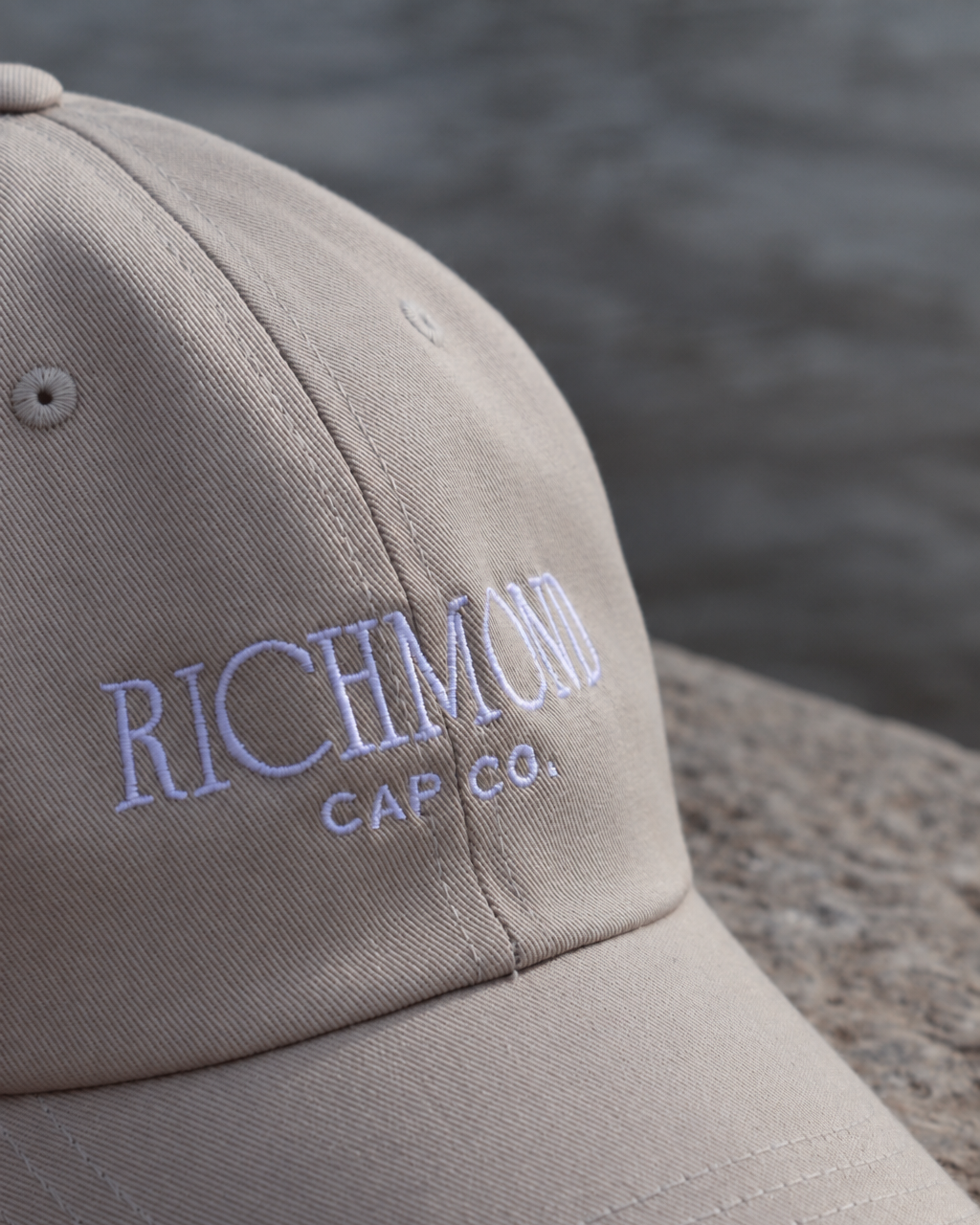 A close-up shot of a beige baseball cap with 'RICHMOND CAP CO.' embroidered in light blue on the front, resting on a stone surface with a blurred water background.