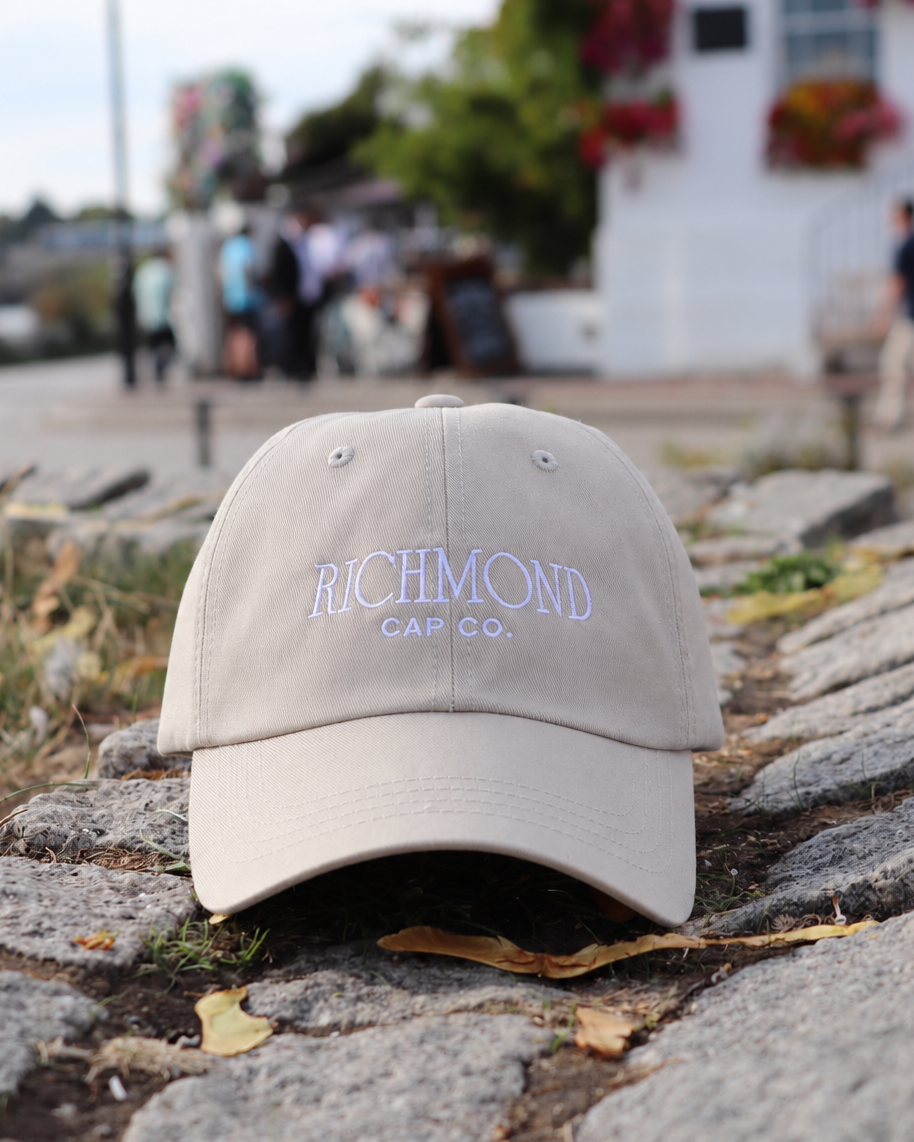 A beige baseball cap with 'RICHMOND CAP CO.' embroidered in light blue on the front, resting on a stone path outdoors with a blurred background of a white building and people.